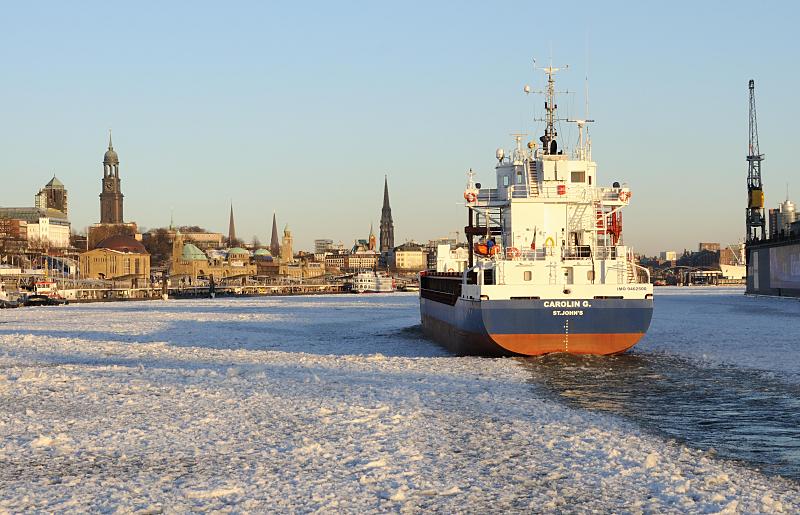 Fotos der Jahreszeiten - Winter in Hamburg, Panorama der Hansestadt; Containerfrachter.  112_5837 |Ein Frachtschiff luft in den Hamburger Hafen ein - das Wasser der Elbe ist mit Treibeis bedeckt. Die Abendsonne scheint auf die Hamburger Silhouette mit den unterschiedlich hohen Trmen. Rechts ein Kran am Schwimmdock der Hamburger Werft Bohm + Voss. www.fotograf-hamburg.de