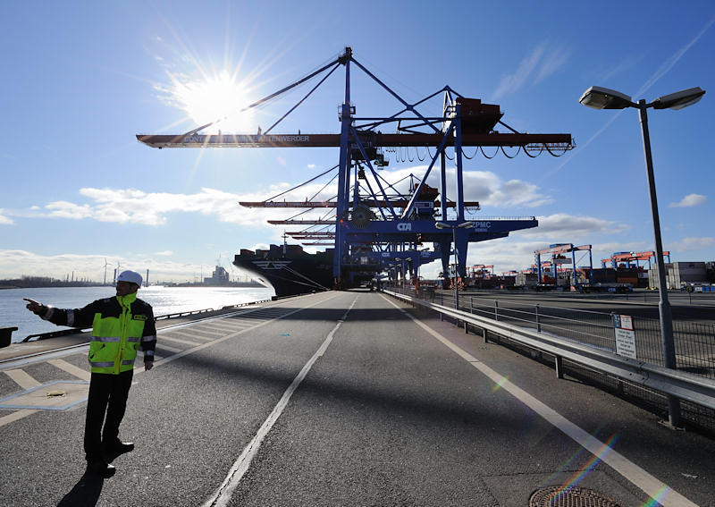 137_0079 | Ein HHLA Mitarbeiter gibt auf der Kaianlage des Container Terminal Altenwerder Anweisungen. Im Hintergrund Containerbrcken und das Containerfrachtschiff HYUNDAI FORCE. Beim hochautomatisierten Containerumschlag auf dem HHLA Container Terminal Altenwerder lschen und laden die mit zwei Katzen ausgestatten Containerbrcken an der Wasserseite die Stahlkisten vom bzw. auf das Schiff.  bilder-hamburg.de