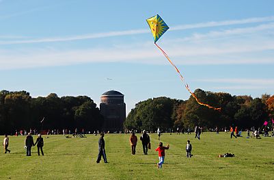 011_25985 | Kinder lassen auf der grossen Wiese im Stadtpark einen Drachen steigen; er hat eine Sonne aufgemalt und einen langen bunten Schwanz; im Hintergrund das Planetarium. 