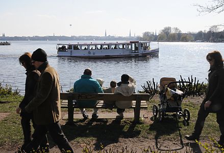 011_25609 | eine Familie sitzt mit ihren Kindern auf einer sonnigen Bank am Alsterufer Hhe Bellevue und geniessen den Ausblick ber die Alster - der Kinderwagen ist neben der Parkbank abgestellt und wirf lange Schatten. Ein Alsterdampfer kommt vom Langen Zug und fhrt weiter Richtung Krugkoppelbrcke. Spaziergnger gehen auf der Wiese entlang der Aussenalster. 