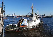 011_26033 | Der Schlepper HANS bei einem Einsatz im Hamburger Hafen; das 26m lange Schiff ist mit einem 1740 PS starken Motor ausgerstet. Das Heck des Schleppers ist mit einer dicken Gummileiste versehen, mit der das Passagierschiff vorsichtig zum Anlegen an den Kai gedrckt wird. Im Hintergrund das Hafenpanorama von Hamburg - ein grosser Frachter luft ein.  www.christoph-bellin.de