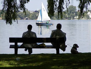 zwei Mnner mit ihrem Hund geniessen die Ruhe auf einer Parkbank am Ufer der Aussenalster mit Blick auf ein Segelboot. 