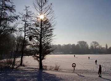 011_25889 | Spaziergnger gehen durch den verschneiten Stadtpark; der Stadtparksee ist zugefroren, ein Rettungsring hngt im Gegenlicht am Seeufer. 