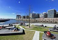 121_1753 | Hamburger und Hamburgerinnen sonnen sich in der Frhlingssonne auf Holzbnken auf den Marco Polo Terrassen in der Hafencity - der Bau in der Mitte des Platzes zeigt noch keine Bltter. Im Hintergrund die Architektur am Dalmannkai des Grasbrookhafens.