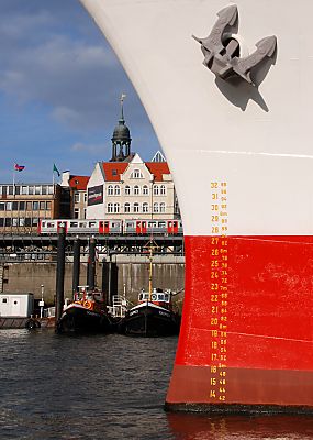 011_25903 | Schiffsbug mit Anzeige fr den Tiefgang des Frachtschiffs und heraufgezogener Anker; im Hintergrund liegen Barkassen und Schlepper im Hamburger Hafen - die Hochbahn fhrt Richtung Haltestelle Landungsbrcken, hinter den Husern ist die Turmspitze vom Hamburger Michel, dem Wahrzeichen der Hansestadt zu erkennen.