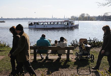 011_25609 | eine Familie sitzt mit ihren Kindern auf einer sonnigen Bank am Alsterufer Höhe Bellevue und geniessen den Ausblick über die Alster - der Kinderwagen ist neben der Parkbank abgestellt und wirf lange Schatten. Ein Alsterdampfer kommt vom Langen Zug und fährt weiter Richtung Krugkoppelbrücke. Spaziergänger gehen auf der Wiese entlang der Aussenalster.