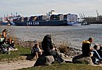 011_26012 |  Hamburger und Hamburgerinnen sitzen in der Frhlingssonne am Elbufer und geniessen den Sonnenschein; sie beobachten den Schiffsverkehr auf dem Fluss: Ein hochbeladener Containerriese fhrt zu seinem Liegeplatz im Hamburger Hafen, wo die Container, die er an Bord hat, gelscht werden. Danach nimmt das Schiff neue Fracht an Bord. www.fotograf-hamburg.de