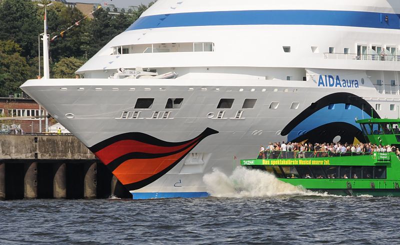 98-4801 |Schiffsbilder aus der Hansestadt Hamburg - Kussmundbug des Kreuzfahrtschiffs Aida Aura, Altona. Das Passagierschiff Aida Aura hat als erstes Kreuzfahrtschiff das Terminal Hamburg-Altona angelaufen. Die Aida Aura wurde 2003 fertig gestellt und hat eine Lnge von 2002 m sowie eine Breite von 28 m. Das Schiff kann 633 Passagiere an Bord nehmen und hat eine Besatzung von 418 Mann. Ein Schiff der Hafenrundfahrt fhrt nahe an den mit einem Kussmund bunt bemalten Bug heran - Hamburgtouristen stehen auf dem Deck und betrachten das Kreuzfahrtschiff.