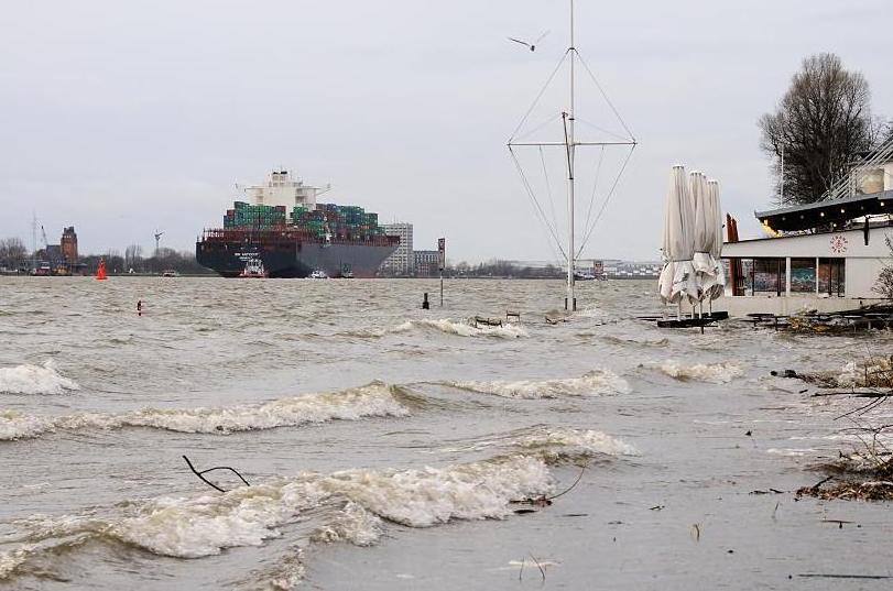 3656_0734 Wellen schlagen auf den Strand bei Hochwasser an der Strandperle in Hamburg Oevelgnne; die Sonnenschirme sind zusammengefaltet, Tische und Sthle gesichert. Ein Containerschiff verlsst den Hamburger Hafen, es wird mit Hilfe von Schleppern in die Fahrrinne gebracht. Der Strand bei der Strandperle und die Uferbefestigung von Oevelgnne stehen bei Sturmflut unter Wasser. 