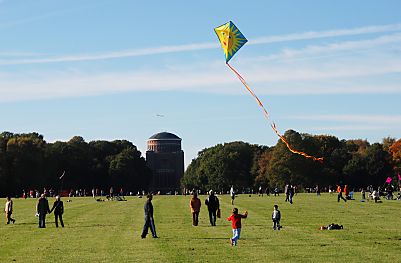 011_25985 | Kinder lassen auf der grossen Wiese im Stadtpark einen Drachen steigen; er hat eine Sonne aufgemalt und einen langen bunten Schwanz; im Hintergrund das Planetarium.